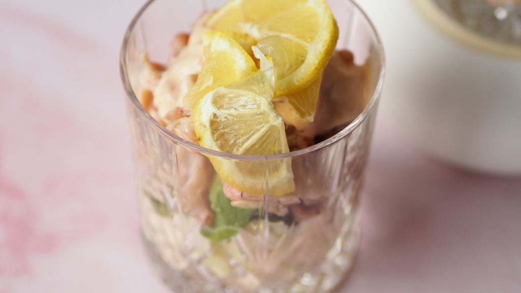 garnalencocktail 2 maal in een kristallen glas met een citroentje tegen een roze, betonnen achtergrond